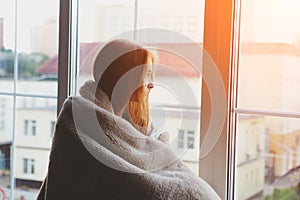 Young woman looking through the window at home wrapped in warm comfy blanket