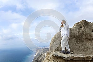 A young woman looks down at the sea while standing on a mountain. Cute blonde in a white jacket. Active recreation in nature