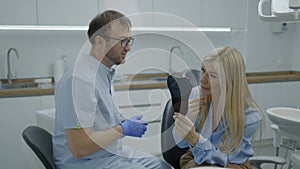 Young woman looks at her smile in mirror while sitting in an armchair at dentist appointment. Male dentist consults