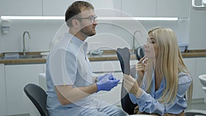 Young woman looks at her smile in mirror while sitting in an armchair at dentist appointment. Male dentist consults