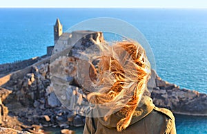 Young woman looks at San Pietro church with sea horizon in background, Lord Byron Natural park in Portovenere village on stone cli