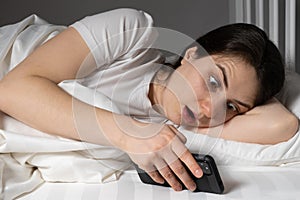 A young woman looks in surprise with her mouth open at the screen of a smartphone lying in bed, a gadget before going to