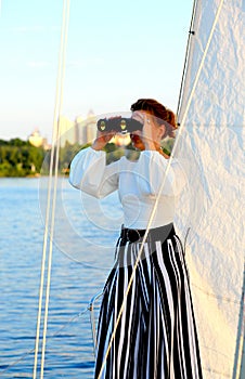 A young woman looks to the left through binoculars while standing on a yacht