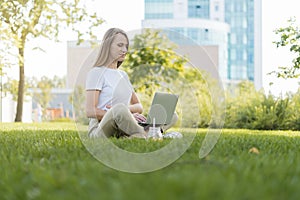 Young woman looks and typing at the laptop, sits on the grass outside in park. Happy and smiling girl working and using