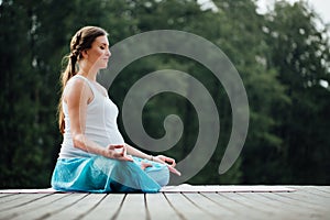 Young woman in the lotus position is practicing yoga in the forest next to the river. sitting on mats the wooden pier.