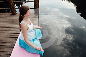 Young woman in the lotus position is practicing yoga in the forest next to the river. sitting on mats the wooden pier.