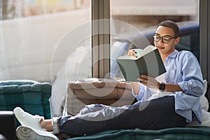 Young woman lounging comfortably on a sofa by the window, lost in an engrossing book