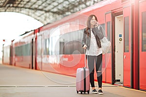 Young woman with luggage talking by cellphone at a train station. Caucasiam tourist waiting her express train while her