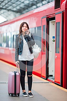 Young woman with luggage talking by cellphone at a train station. Caucasiam tourist waiting her express train while her