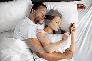 Young woman lying on bed in arms of her black husband and using mobile phone to text her lover, top view