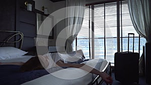 Young woman is lying on bed in hotel room. Lady is tired after travel and resting in room with large window.