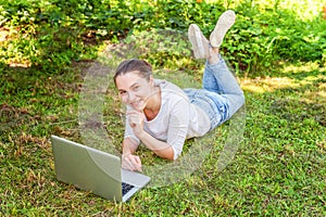 Young woman lying on green grass lawn in city park working on laptop pc computer. Freelance business concept