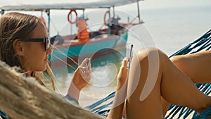 Young woman lying on hammock with smart phone and bank card at the sandy beach