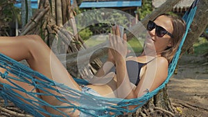 Young woman lying on hammock with smart phone at the sandy beach in slow motion