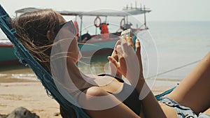 Young woman lying on hammock with smart phone at the sandy beach in slow motion