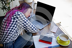 Young woman lying on her arms on the table in cafe in front of laptop with cup of coffee, sleepy.