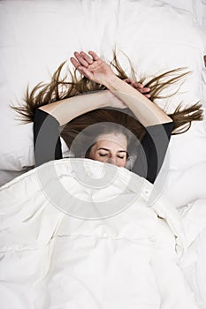 Young woman is lying in her bed with closed eyes, smiling under her blanket after a restful sleep.