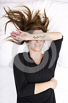 Young woman is lying in her bed, joking. She is replacing her eyes by covering it with her painted arms. photo