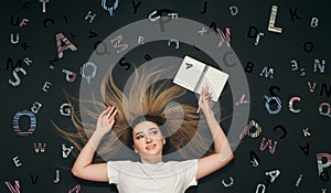 Young woman lying with pencil and notebook top view on black studio background with scattered letters written and chalk shaded on