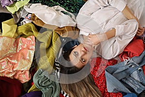 Young woman lying on pile of clothes, top view. Fast fashion concept