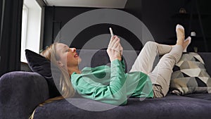 Young woman lying on sofa chatting typing message using smart phone, posting stories on social media