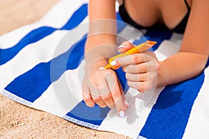 Young woman is lying on striped towel on the sand at the beach and applying sun cream on her hand