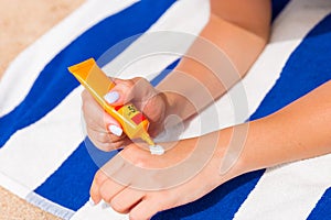 Young woman is lying on striped towel on the sand at the beach and applying sun cream on her hand