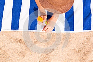 Young woman is lying on striped towel on the sand at the beach and applying sun cream on her hand