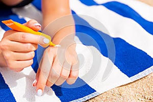 Young woman is lying on striped towel on the sand at the beach and applying sun cream on her hand