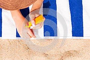 Young woman is lying on striped towel on the sand at the beach and applying sun cream on her hand