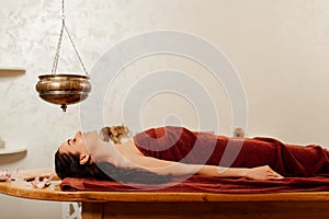 Young woman lying under shirodhara vessel during ayurvedic procedure