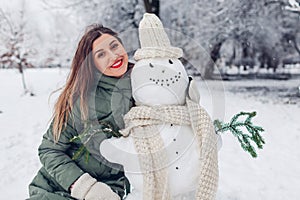 Young woman made snowman outdoors in snowy winter park, dressed it in hat, scarf and attached fir branches in hands