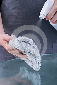 Young woman maid in apron is cleaning, wiping down office glass table surface with spray bottle cleaner, wet rag, close up,