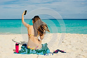 Young woman in a swimsuit in the sand makes selfie on the beach on her smartphone