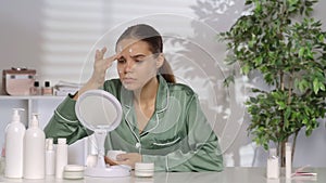 A young woman without makeup applying white cream on problem skin areas of the face. A woman in green silk pajamas