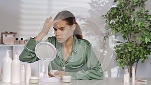 A young woman without makeup applying white cream on problem skin areas of the face. A woman in green silk pajamas