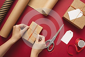 Young woman making bow on kraft gift boxe with orange thread on a red background.