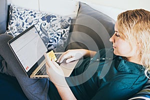 Young woman making online purchase with credit card on the sofa, looking at laptop screen and smiling