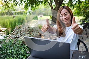 A young woman making and showing thumbs up hand sign while working on laptop in the outdoors