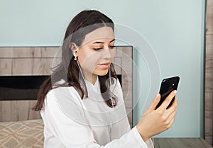 Young woman is making a video call on her smartphone. In room. There is a earphones in the woman`s ears