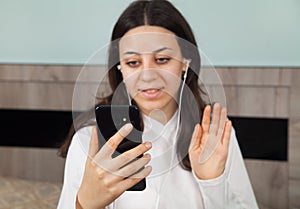 Young woman is making a video call on her smartphone. In room. There is a earphones in the woman`s ears