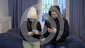 Young woman and man in jackets are sitting on the bed with phones in their hands. A young family has not paid their