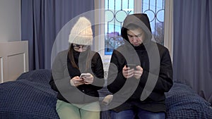 Young woman and man in jackets are sitting on the bed with phones in their hands. A young family has not paid their