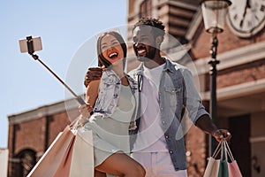 Young woman and man with shopping bags and taking selfie with smartphone using selfiestick on the street