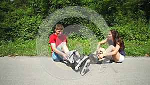 Young woman and man sitting on track, putting their rollerblades on their feet.