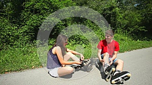 Young woman and man sitting on track, putting their rollerblades on their feet.