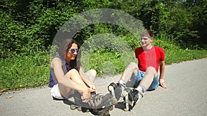 Young woman and man sitting on track, putting their rollerblades on their feet.