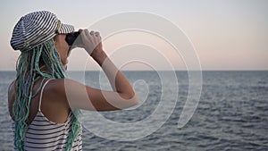 A young woman in a marine striped dress looks through binoculars at the sea or ocean