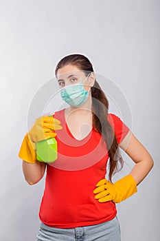 A young woman in a mask and yellow gloves holds a green spray bottle in her hands, isolated on a white background.