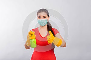 A young woman in a mask and yellow gloves holds a green spray bottle in her hands,  on a white background.